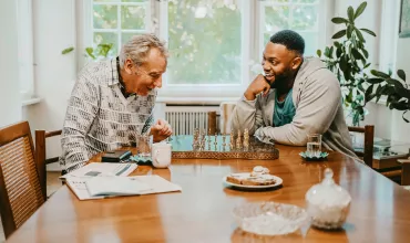 Two men, seated indoors around a table, engaged in a conversation and playing chess Two men, seated indoors around a table, engaged in a conversation and playing chess