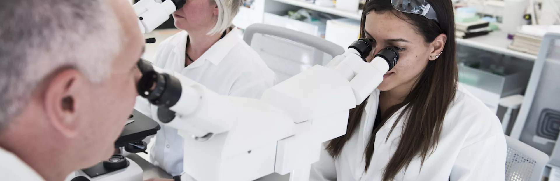 Foto dentro de um laboratório com duas mulheres olhando através de um microscópio. As mulheres vestem jaleco branco e usam óculos de proteção acima da cabeça. A mulher na frente tem cabelos longos castanhos. 