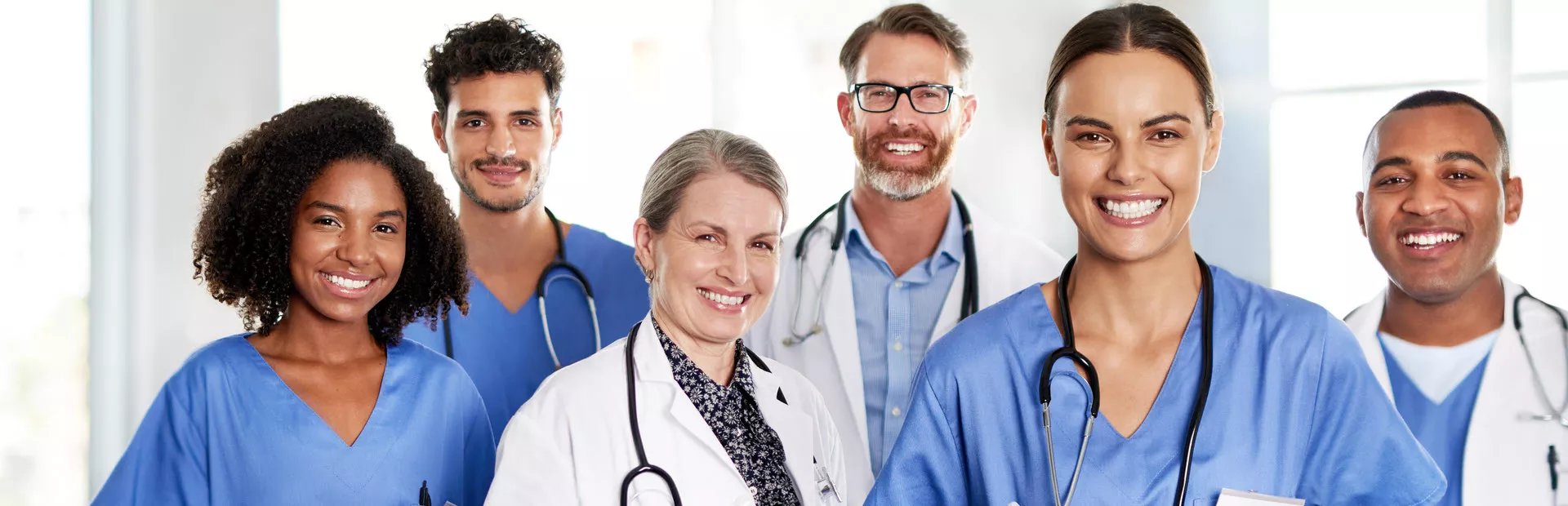 Foto de uma equipe hospitalar de 5 pessoas sorrindo para a câmera. Da direita para esquerda, uma mulher negra de cabelo médio crespo com uniforme azul, um homem pardo de barba e bigode escuros e uniforme azul, uma mulher branca com cabelo preso e jaleco branco, um homem branco de barca marrom usando óculos e jaleco branco, uma mulher de pele média usando uniforme azul.  Todos usam estetoscópio em volta do pescoço.