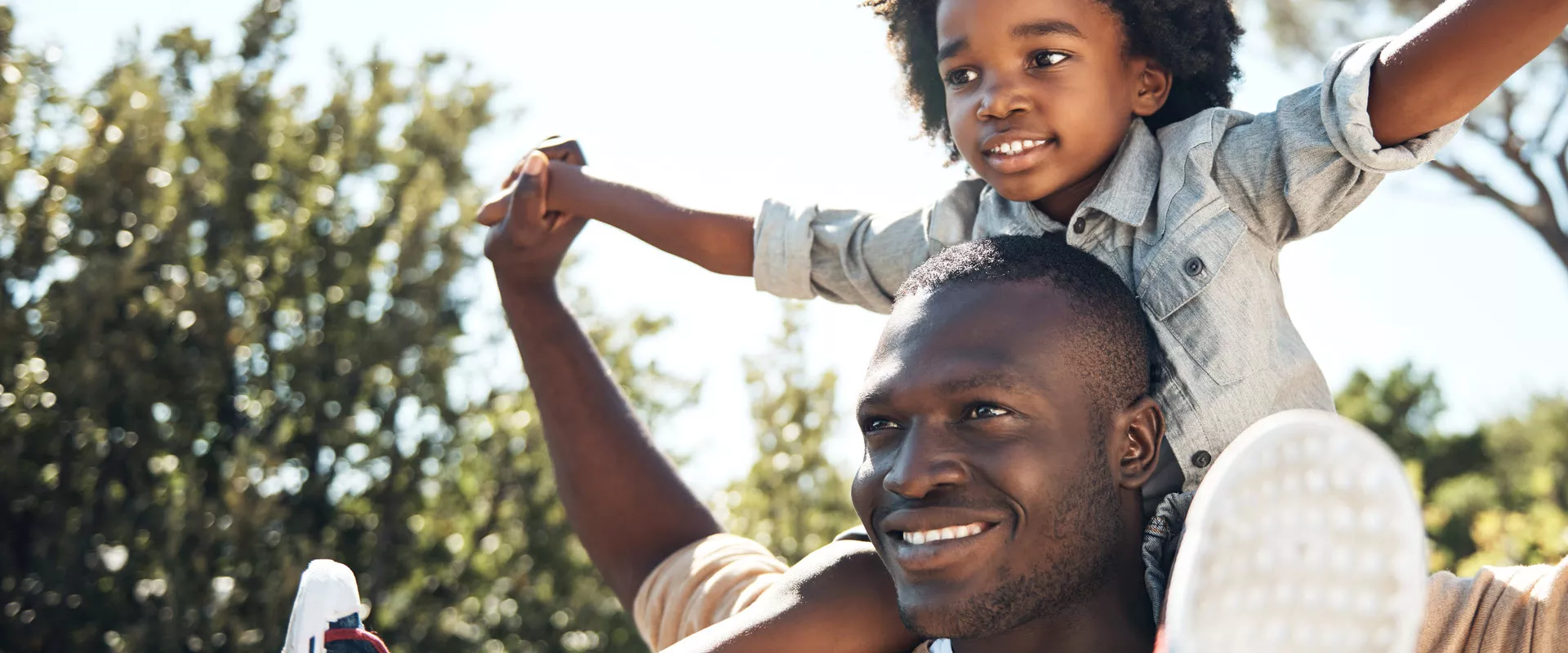 Foto de um homem sorrindo com uma criança sentada sob os seus ombros de braços erguidos para o ar, ao fundo da imagem aparece o céu e os galhos de árvores, com folhas. O homem é adulto, preto, de cabelo curto. A criança é preta, tem cabelos cacheados médio escuro, usa uma blusa social e calça jeans. Ambos olham para o horizonte num dia ensolarado. 