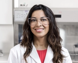 A female Novartis scientist wearing a white lab coat and glasses, smiles in front of laboratory equipment.